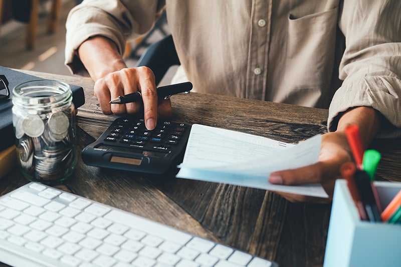 A person using a calculator with a paper in hand an a jar of coins off to the side.